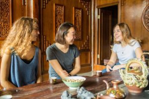 Three women talk and laugh at a carved wooden tea table
