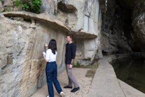 Two people look at carvings on a cliff wall beside a cave entrance