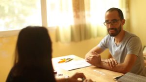 A smiling man in glasses talks across a sunlit table during a lesson