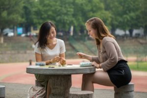 Two women play a board game at a round stone table outdoors