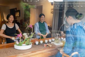 Two smiling women prepare tea at a long table, seen through a window