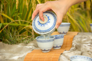 Tea is poured from a lidded cup into small cups beside rice stalks