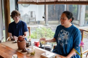A tea host pours tea for a man at a wooden table by a window