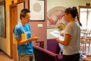 A man in a blue T-shirt gestures while talking with a woman in a lounge
