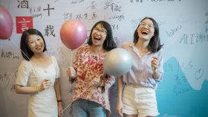 Three women laugh with balloons in front of a signed CLI wall