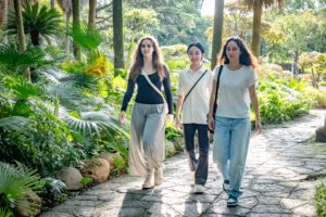 Three women walk along a stone path through a lush garden