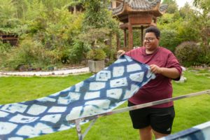 A man holds up a blue tie-dye cloth on a garden lawn