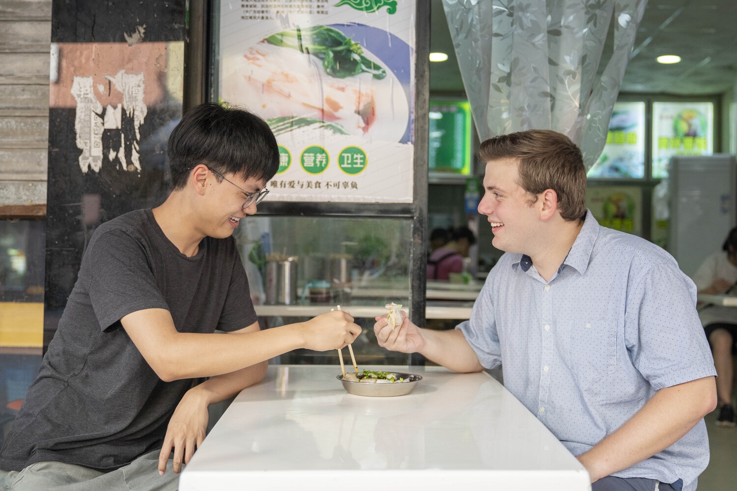 Two men sit across from each other eating street food at a small table.