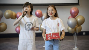 Two women stand by balloons, one with a microphone and one with a CLI box