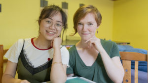 Two smiling women pose close together in a yellow room