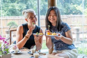 Two smiling women hold plates of snacks by a window onto a garden