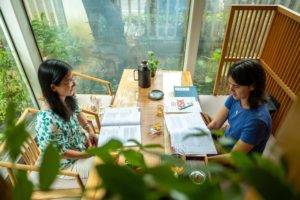 Two women study at a wooden table in a sunlit glass room