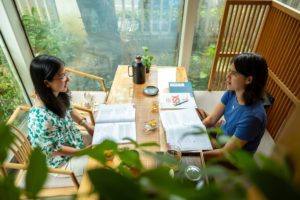 Two women study with textbooks at a wooden table by large windows