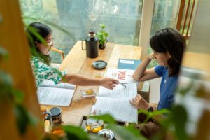 Two women study at a wooden table, seen through leafy plants