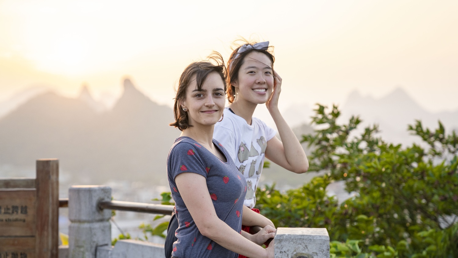 Two women standing together at a mountain overlook during sunset with karst peaks behind them.