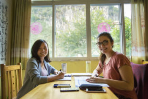 Two women smile at a table with notebooks in a bright room