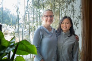 Two women smile side by side in front of a bright window