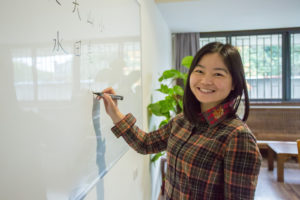 A smiling teacher writes on a whiteboard with a marker