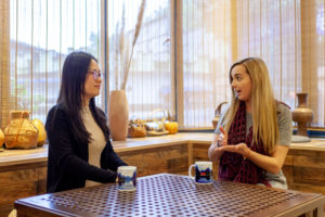Two women talk across a table in front of a large window