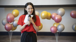 A woman holds a microphone in front of a wall of balloons