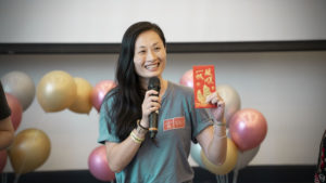 A woman with a microphone holds up a red envelope in front of party balloons