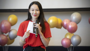 A woman in red holds a microphone and a prize slip in front of balloons