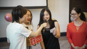 Three women laugh as one speaks into a microphone beside a CLI prize box