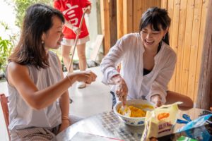 Two women smile while serving food from a pot at a table