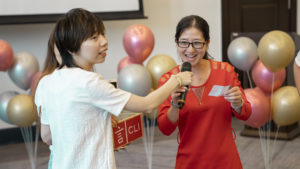 A woman in white holds a microphone for a woman in red reading a prize slip