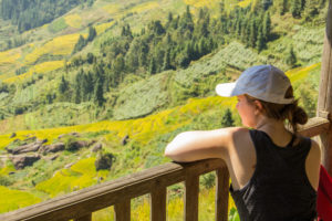 A woman in a cap leans on a wooden railing above bright rice terraces