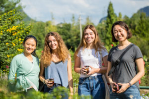 Four women stand outdoors holding cups and smiling with mountains behind