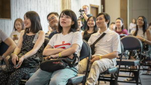 An audience sits on folding chairs listening to an indoor presentation