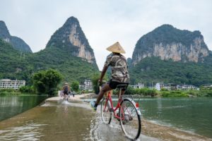 A cyclist in a conical hat rides through water over a weir beneath karst peaks