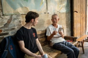 A man in a black T-shirt chats with a man holding a phone on a bench