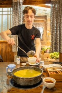 A man in a black T-shirt ladles yellow broth at a hot pot table