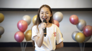 A woman in a white 言 T-shirt speaks into a microphone in front of balloons
