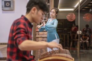 A guzheng player performs while a dancer in blue moves behind him
