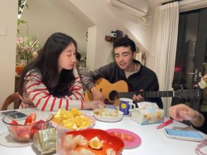 A man plays guitar beside a woman at a table of fruit at home