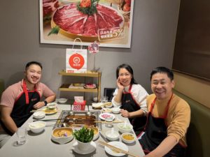 Three people in aprons smile around a hot pot table in a restaurant