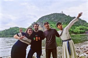 Four friends pose and wave on a riverbank below a green hill