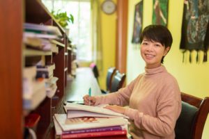 A smiling woman in a pink sweater writes at a desk beside bookshelves