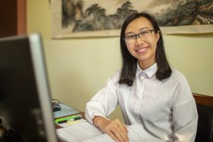 A smiling woman in a white shirt sits at a desk beside a computer