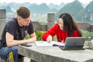 A teacher and student study with a laptop on a stone table above karst peaks