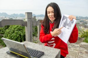 A teacher in a red hoodie shows a printed page to a hilltop laptop