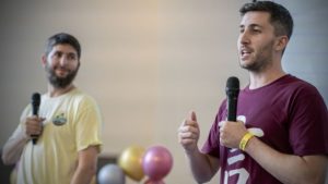 Two men with microphones speak on stage in front of balloons