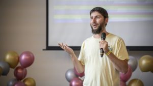 A man gestures while speaking into a microphone in front of balloons