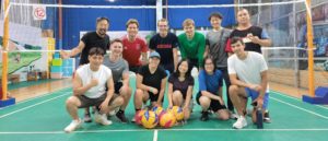 A group kneels and stands behind a row of volleyballs on an indoor court