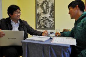 Two men go over papers at a desk with a laptop