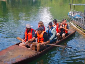People in life jackets sit in a narrow wooden boat beside a dock