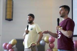 Two men with microphones, one gesturing, address a party in front of balloons